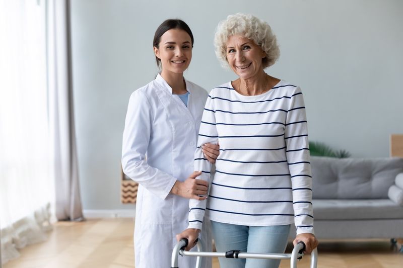 أخصائية العلاج الطبيعي التي تقدم الرعاية تساعد مريضة عجوز سعيدة على الوقوف مع المشاية.'