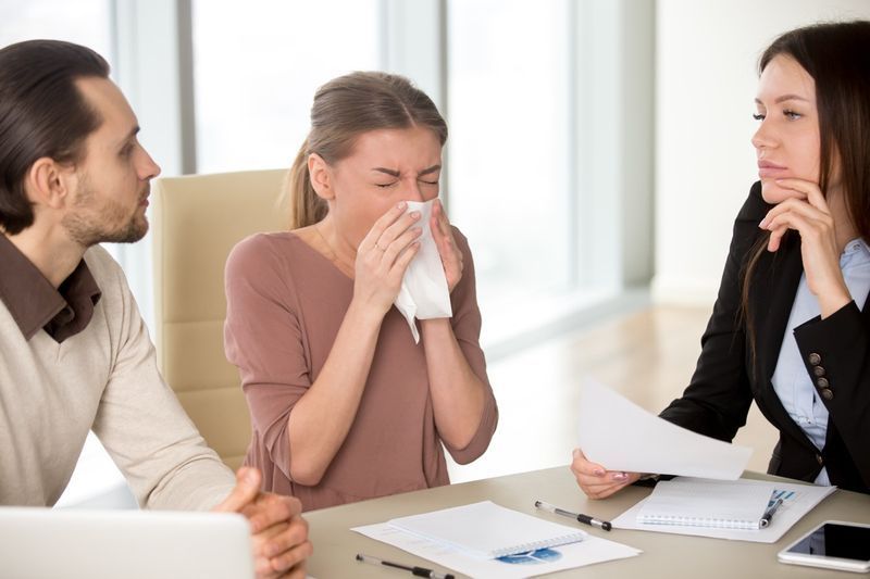 امرأة ذات منديل تعاني من نوبة عطس ، وتنفخ أنفها أثناء العمل مع زملائها في الاجتماع ، وأصيبت بالبرد ، وأعراض الأنفلونزا'