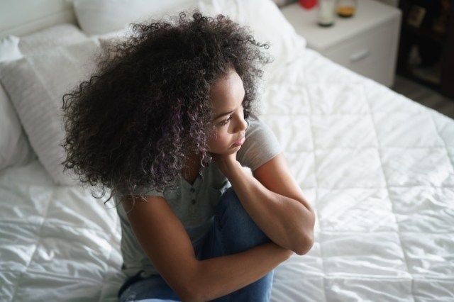امرأة شابة وحيدة تجلس على السرير. فتاة من أصل اسباني مكتئبة في المنزل ، تنظر بعيدًا بتعبير حزين'