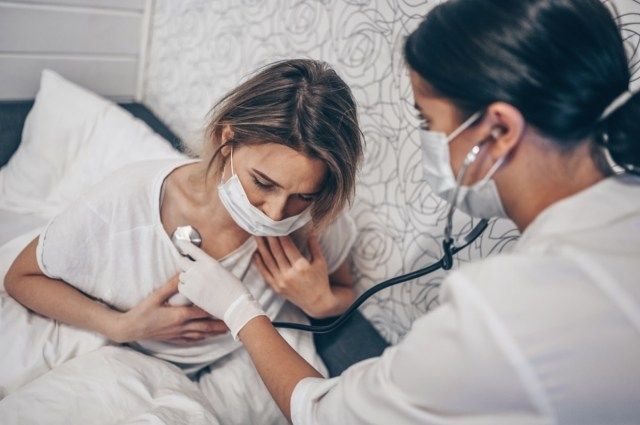 ممرضة طبية ترتدي قناعًا واقيًا للوجه تستمع إلى التنفس بواسطة سماعة الطبيب للاشتباه في فيروس كورونا (COVID-19).'