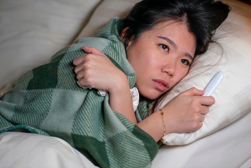 امرأة ترقد على سريرها في المنزل مريضة تعاني من نزلة برد ودرجة حرارة مغطاة ببطانية وتشعر بتوعك وارتفاع في درجة الحرارة'