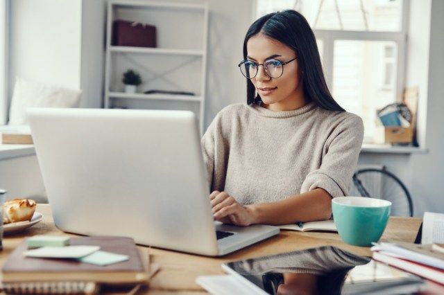 امرأة في ملابس غير رسمية تستخدم الكمبيوتر المحمول وتبتسم أثناء العمل في الداخل'
