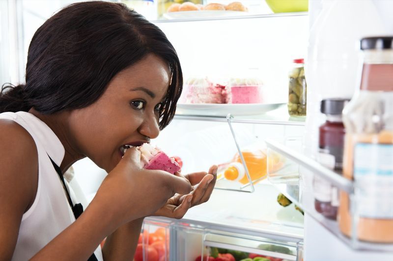 امرأة أفريقية تأكل شريحة من الكعكة بالقرب من الثلاجة المفتوحة'