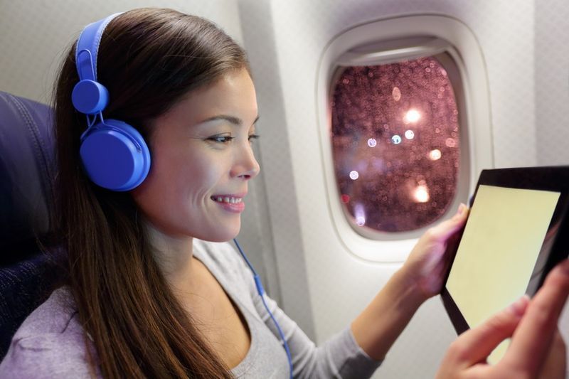 الراكب في الطائرة باستخدام الكمبيوتر اللوحي. امرأة في مقصورة الطائرة تستخدم جهازًا ذكيًا تستمع إلى الموسيقى على سماعات الرأس'