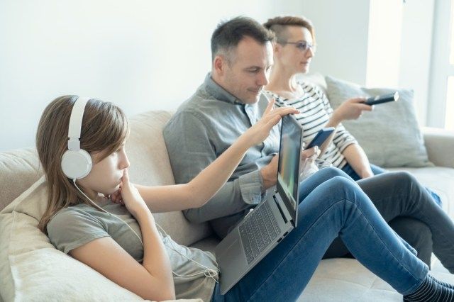 فتاة صغيرة تستخدم الكمبيوتر المحمول على السرير بينما يشاهد الأب والأم التلفزيون ولا ينتبهان لها'