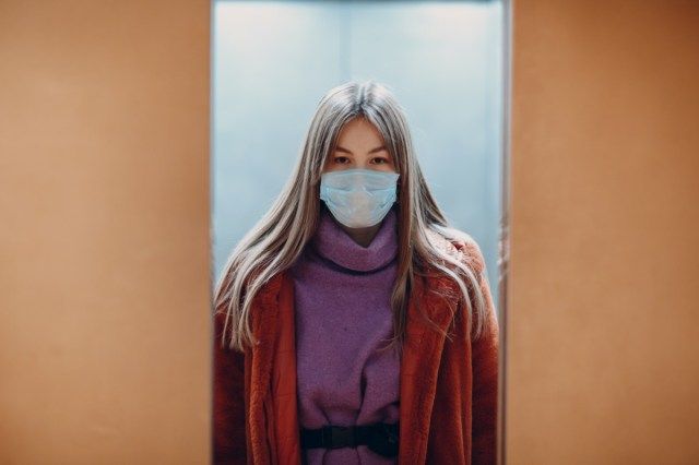 جرو، صيني، وقوف المرأة، في المصعد، إلى داخل، القناع الطبي. الأبواب تغلق. مفهوم جائحة فيروس كورونا COVID-19.'