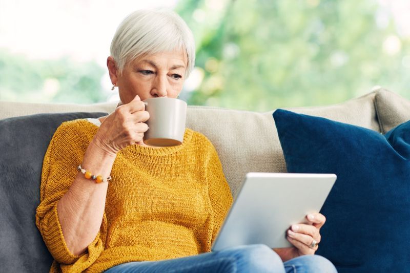 امرأة مسنة تستخدم جهازًا لوحيًا رقميًا وتناول القهوة على الأريكة في المنزل.'