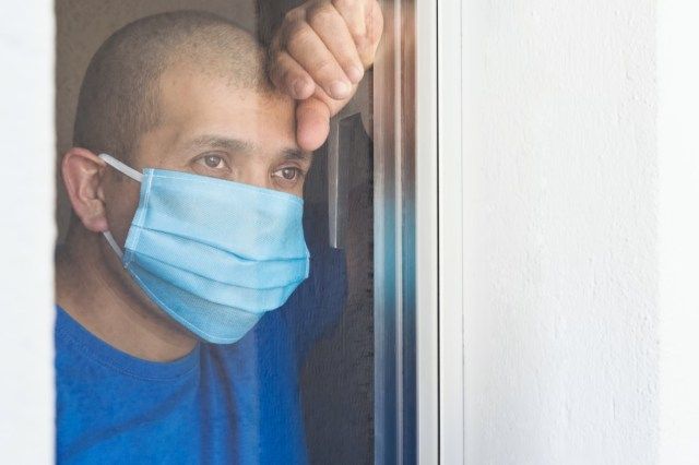 رجل مريض يرتدي قناع وجه ينظر من النافذة ويتم عزله في المنزل'