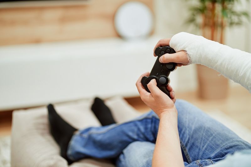 رجل ذو ذراع مكسورة في قالب من الجبس يمسك جهاز التحكم ويلعب في لعبة فيديو أمام التلفزيون.'