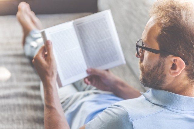 رجل يرتدي ملابس غير رسمية ونظارات يقرأ كتابًا بينما يرقد على الأريكة في المنزل'