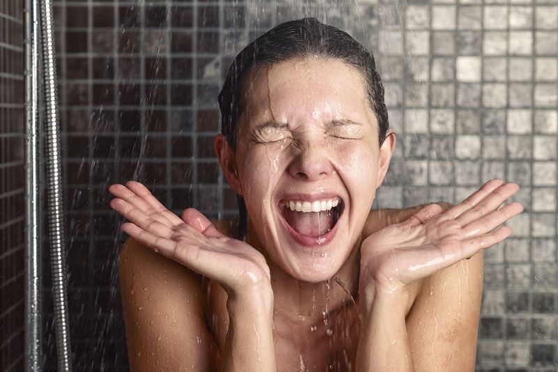 امرأة شابة تتفاعل بصدمة تجاه ماء الدش الساخن أو البارد وهي تقف تحت رأس الدش تغسل عينيها من شعرها ويداها مرفوعتان وفمها مفتوح'