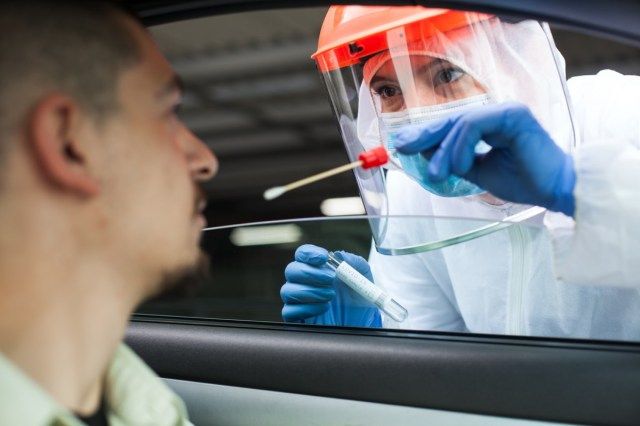 عامل طبي يجري اختبار COVID-19 من السيارة ، ويأخذ عينة مسحة أنف من مريض ذكر من خلال نافذة السيارة.'