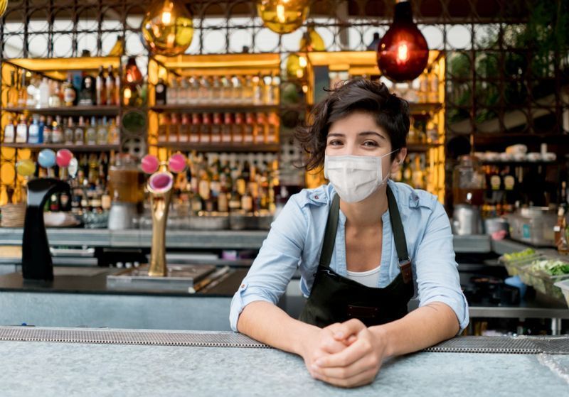 صورة لنادلة سعيدة تعمل في مطعم ترتدي قناع الوجه.'
