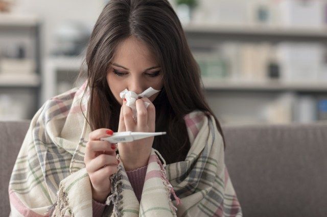 امرأة شابة مريضة في المنزل على الأريكة ، تغطى ببطانية ، وتقاس درجة الحرارة وتنفخ أنفها بمنديل ورقي'