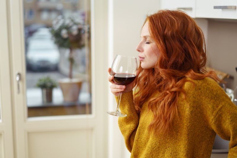 امرأة شابة حمراء الشعر تقف في شقتها وهي تحتسي كأسا من النبيذ الأحمر وعيناها مغمضتان بسرور'