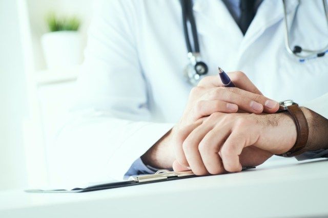 طبيب رجل يحمل سماعة طبية ينظر إلى ساعة اليد. طبيب يرتدي الزي الأبيض على خلفية المكتب ينتظر المرضى'