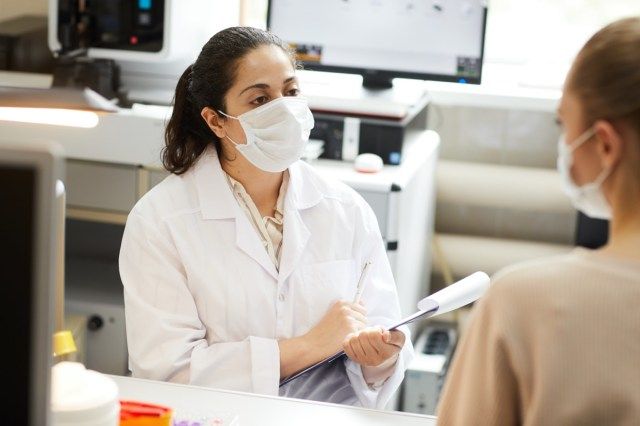 طبيبة ترتدي قناعًا تدون الملاحظات في البطاقة الطبية أثناء التحدث إلى المريض في المستشفى'