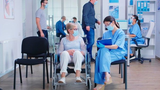 ممرضة ترتدي قناع وجه ضد فيروس كورونا وتدون ملاحظات على الحافظة أثناء التحدث مع امرأة مسنة على كرسي متحرك'