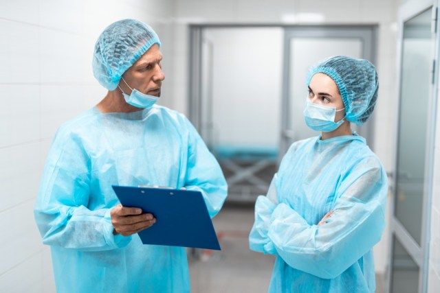 طبيبان محترفان يرتديان الزي الطبي الأزرق يقفان أمام بعضهما البعض في ممر المستشفى ويبدو أنهما مدروسان'