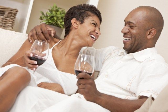 زوجان يجلسان في المنزل معًا يضحكان ويشربان كؤوس من النبيذ الأحمر'