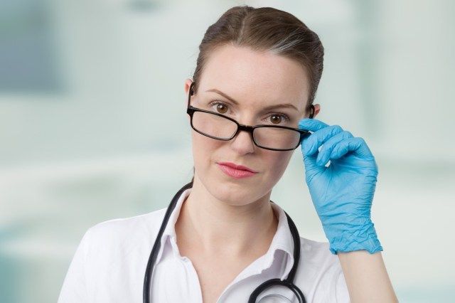 طبيبة ترتدي قفازات طبية زرقاء وسماعة طبية تنظر فوق نظارتها أمام غرفة العيادة'