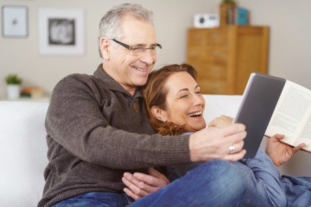 زوجان حنون في منتصف العمر يسترخيان على أريكة معًا في المنزل ويضحكان على شيء ما على الكمبيوتر اللوحي ، طبيعي وتلقائي'