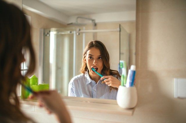 صورة لامرأة جميلة تفريش أسنانها وتنظر في المرآة في الحمام.'