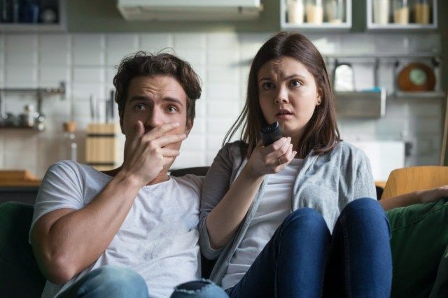 زوجان خائفان من جيل الألفية يشاهدان فيلم رعب على شاشة التلفزيون ويمسكان جهاز التحكم عن بعد في المنزل'