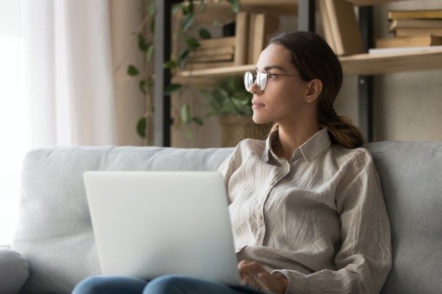 امرأة تجلس على الأريكة ممسكة بالكمبيوتر المحمول تبدو بعيدة تفكر في تشتت انتباهها عن العمل عبر الإنترنت'