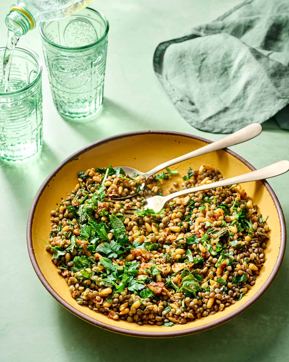 Spoon and fork in a big bowl of lentil salad on green table.