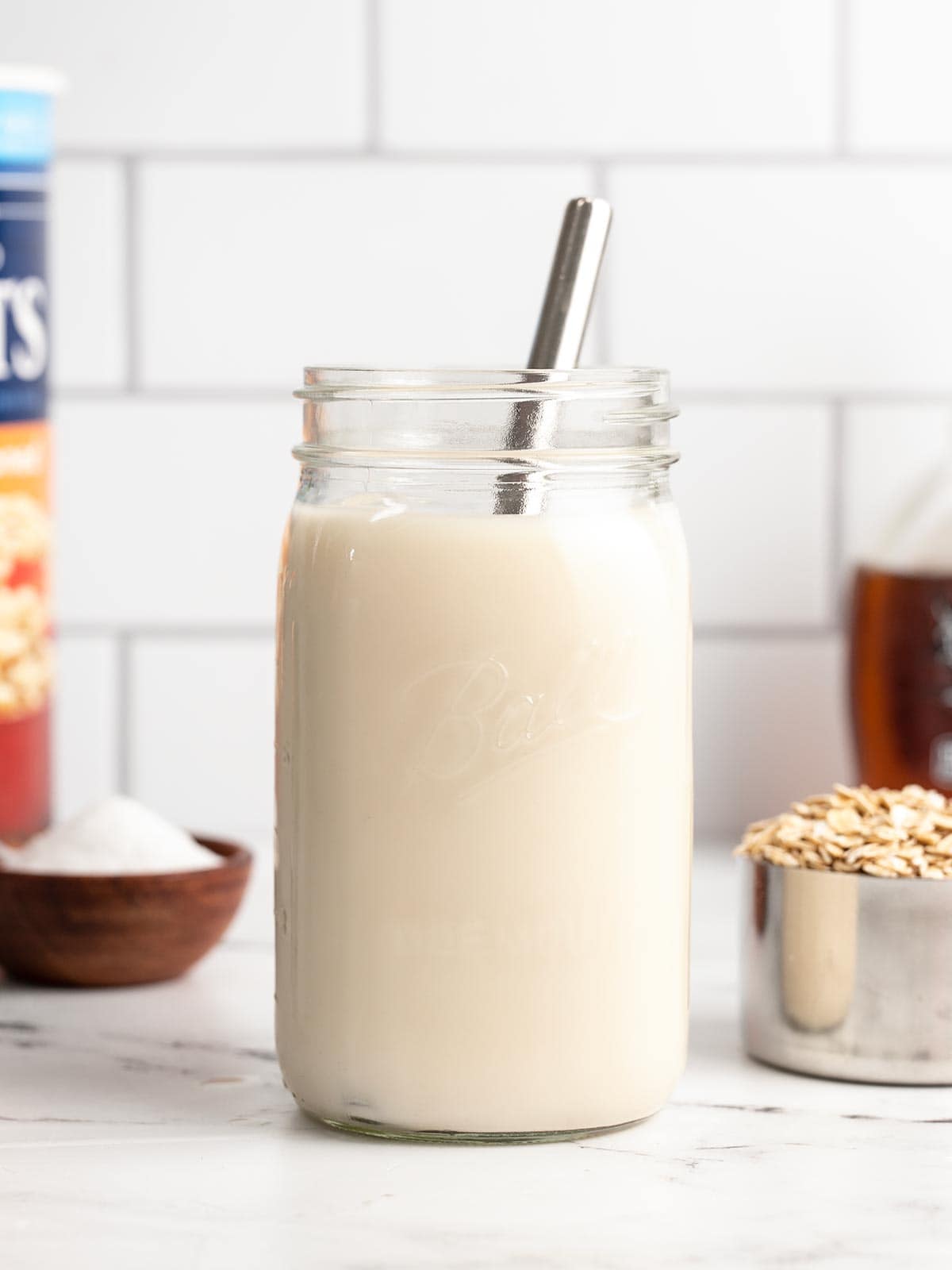 A jar of oat milk with a metal straw and ingredients in the background.