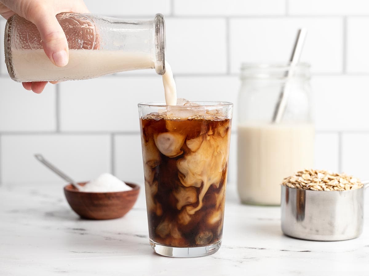 Oat milk being poured into an iced coffee from a glass carafe.