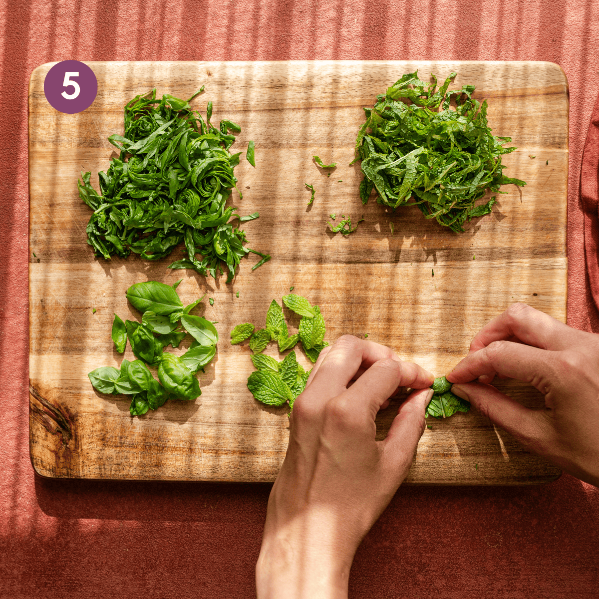 thinly sliced basil and mint on a cutting board with woman's hands folding up mint leaves to chiffonade.