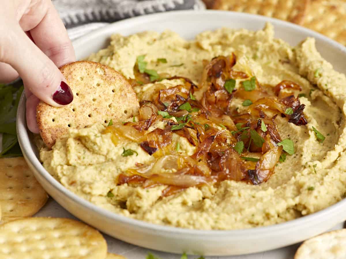 Side view of a cracker being dipped into a bowl of chickpea spread with caramelized onions.